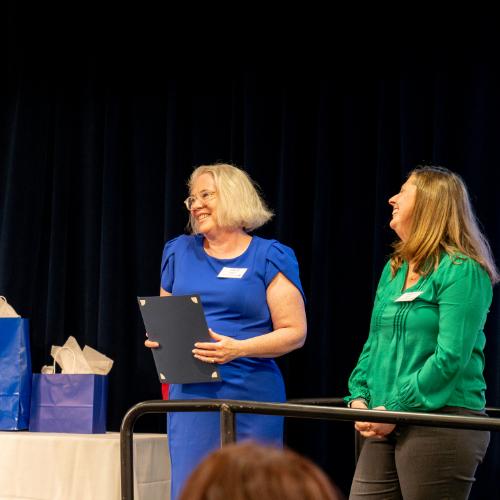 two women laugh as they stand on stage; Chelsea Ridge in green and Kathy Agee in blue dress