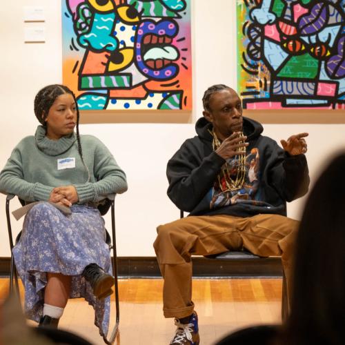 two people sitting in chairs answering questions from audience in art gallery; brightly colored artwork is on the walls behind them