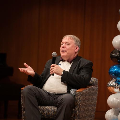 Andy Beachnau, seated in chair with microphone, gestures with hand. A balloon arch is at his right