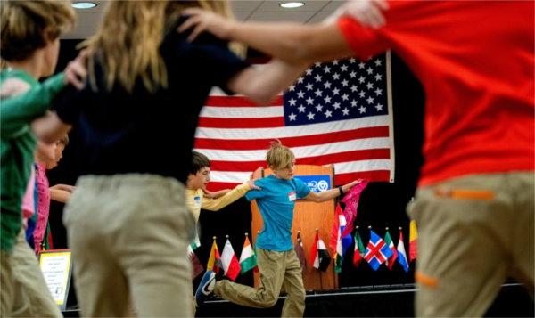  Middle-school age children hold onto each other's shoulders while dancing in a circle with an American flag behind them. 
