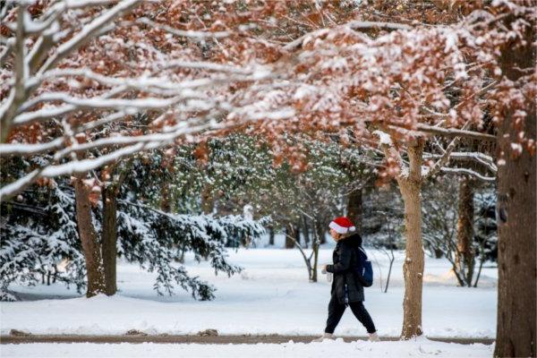 一个人 walks on a sidewalk in snowy surroundings wearing a Santa hat.  