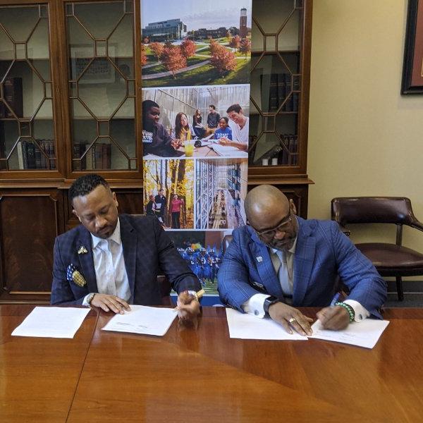 B. Donta Truss, left, and Kevin James sign papers while seated at a table; a banner from GVSU is behind them