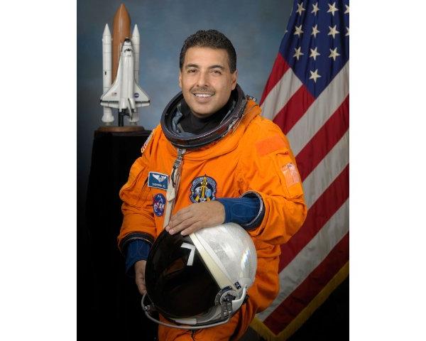 一个穿着橙色制服，拿着宇航员头盔的人微笑着. 背景是一面美国国旗和一个火箭模型.