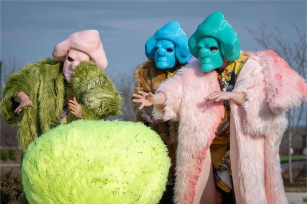 People wearing furry coats of pink, gold and green and wearing masks of green, blue and pink use their hands to gesture while standing in front of a green, furry sphere.