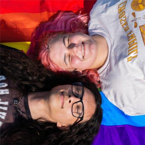 Two people - one with pink hair, 另一个留着黑色长发，戴着眼镜，躺在彩虹骄傲旗上，对着镜头咧嘴笑着.