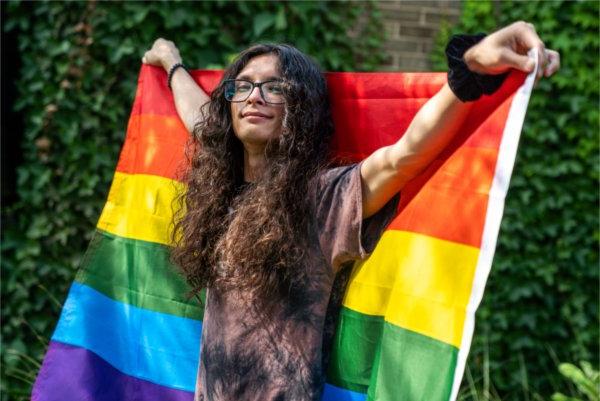 Lex Larkspur, a person with long wavy black hair and a brown shirt, holds a rainbow pride flag behind her back.