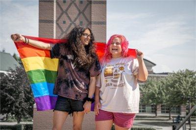 Two people, one with pink hair and the other with long black hair and glasses, hold a pride flag behind them and stand in front of the Cook Carillon clock tower.
