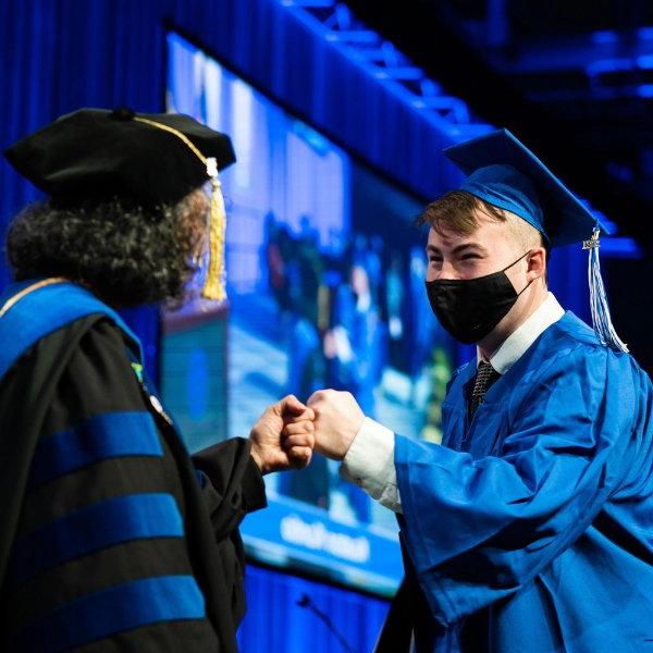 A graduate in a cap and gown fist bumps with a dean.