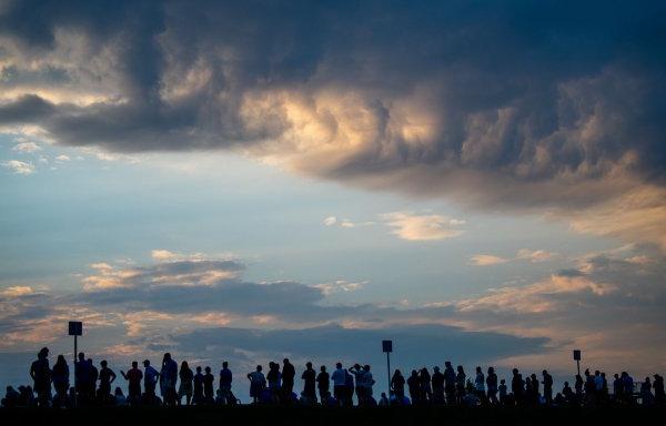 Silhouetted people stand underneath a cloudy evening sunset during a football game.  