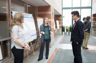 A Student Scholars Day participant stands next to a poster explaining it to another person. A person in a white shirt looks on at right.