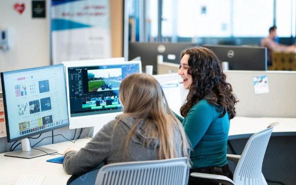 Two female students sit and utilize the dual monitor setups in the Digital Creator Lab. 