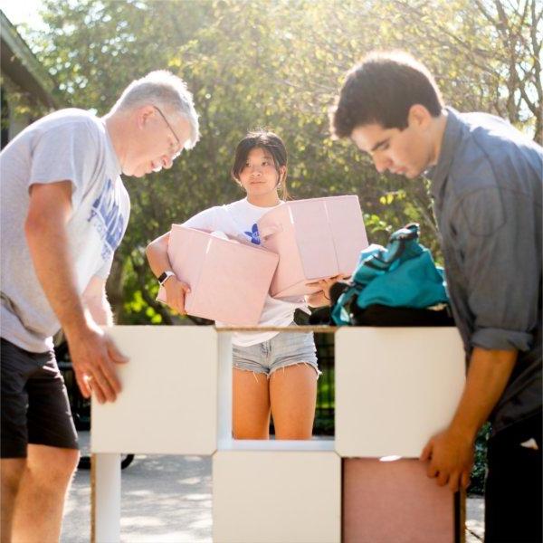 Two people stand on either side of a multi-tiered storage container while another person carrying bins stands behind them.