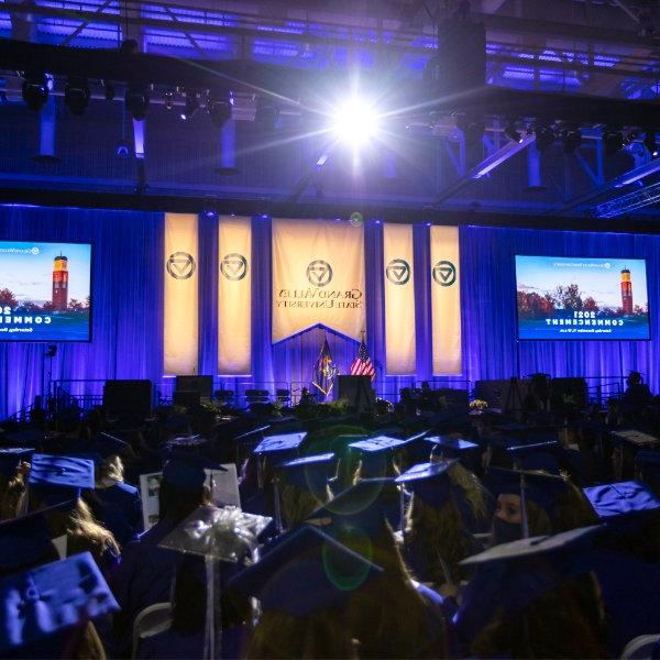 A stage with Grand Valley banners hanging over a blue curtain. A large crowd of graduates in graduation caps and gowns.