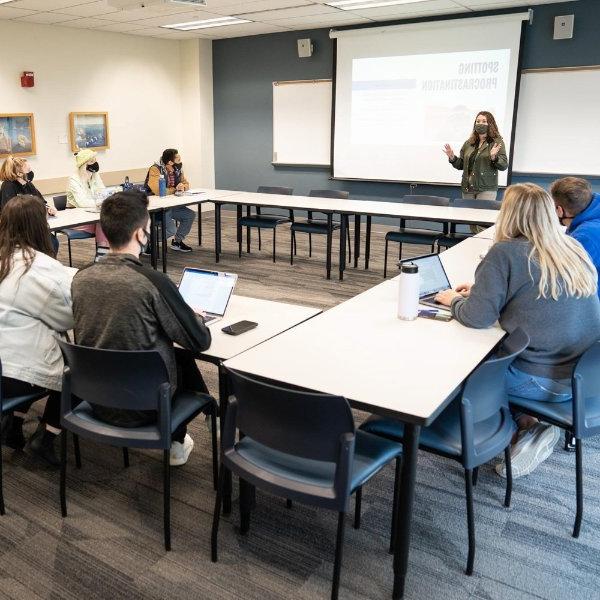 杰西卡·菲尔莫尔(Jessica Fillmore)领导着一个小组研讨会, 站在教室前面讲话, 学生们坐在盒子形状的桌子旁的椅子上. 发现拖延症