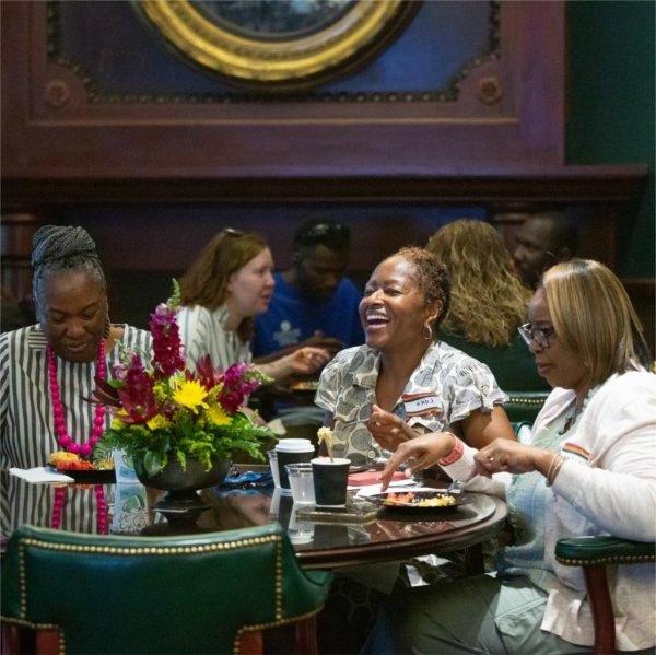 Three well-dressed women with name tags on sit at a table and laugh together as they eat breakfast.